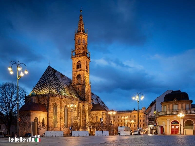 Abendliche Ansicht des Bozner Doms mit seinem farbigen Ziegeldach und gotischem Turm, umgeben von stimmungsvollem Licht und historischen Gebäuden.