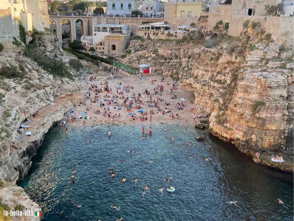Blick auf die belebte Bucht von Lama Monachile in Polignano a Mare, umgeben von Klippen und historischen Gebäuden, mit zahlreichen Badegästen im Wasser und auf dem Strand.
