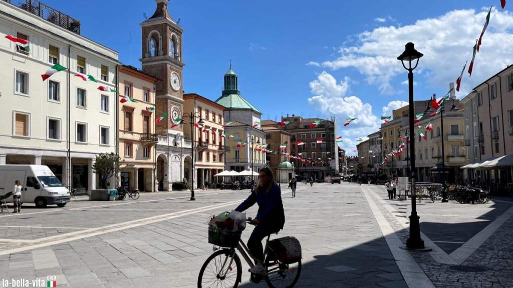 Uma mulher anda de bicicleta numa rua movimentada de uma cidade com edifícios históricos e bandeiras italianas, sob um céu azul claro.